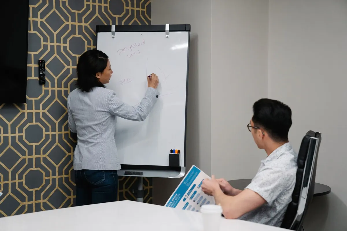 Two colleagues engaged in a business discussion with notes on a whiteboard during a meeting.